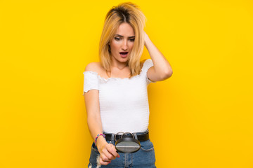 Young blonde woman over isolated yellow wall holding a magnifying glass