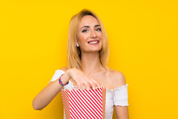 Young blonde woman over isolated yellow wall holding a bowl of popcorns