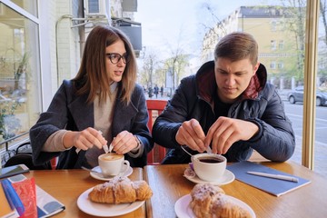 Young male and female friends students sitting in outdoor cafe, talking, drinking coffee, tea, eating croissants. On table textbooks, notebooks, city background.