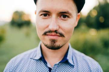 Fototapeta premium Closeup lifestyle portrait of handsome kazakh man posing in summer outdoor on abstract nature background. Smiling happy male with mustache and beard wearing cap. Charismatic adult guy in blue t-shirt.