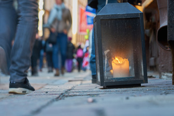 Obraz premium Big metal dirty lantern with burning candle inside, outdoor on a ground with blurred crowded street on a background.