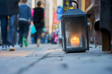  Big metal dirty lantern with burning candle inside, outdoor on a ground with blurred crowded street on a background.