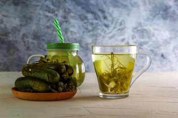Glass cup and a jar of cucumber pickle along with pickled cucumbers on a gray wooden table. Copy space