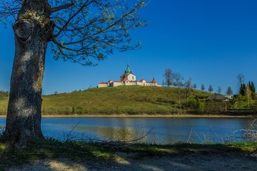 Obraz premium Spring view of Pilgrimage Church of St John of Nepomuk at Zelena Hora on top of a green hill at Zdar nad Sazavou, bright sunny day with clear blue sky, Convent lake, tree and sandy land in foreground