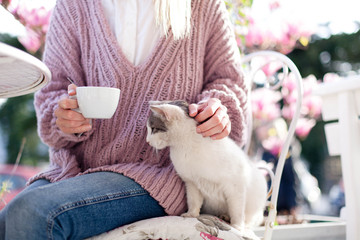Young woman is drinking coffee and stroking cat in spring cafe on city streets. Girl is sitting...