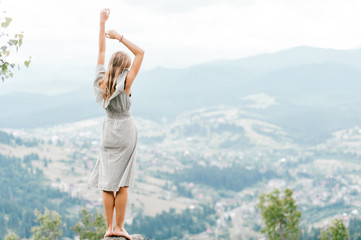 Fototapeta na wymiar Young beautiful barefoot blonde girl with long hair in summer dress standing on top of conquered mountain at stone and enjoying fabulous landscape scenic view with mountains and village in valley