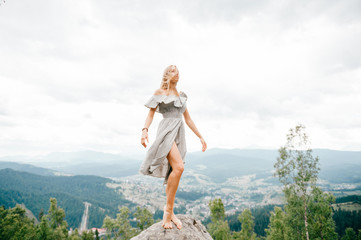 Young beautiful barefoot blonde girl with long hair in summer dress standing on top of conquered mountain at stone and enjoying fabulous landscape scenic view with mountains and village in valley