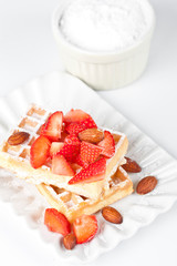 Belgium waffers with sugar powder, strawberries and almonds on ceramic plate on white table.