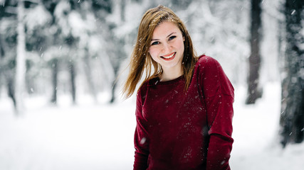 the girl dressed in a maroon sweater stands against the tree trunk against a backdrop of snow-covered winter forest