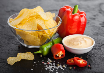 Glass bowl plate with potato crisps chips with onion flavour on black stone table background. Red and green chilli peppers with paprika and hot spicy sauce.