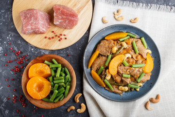 Fried pork with peaches, cashew and green beans on a black concrete background, close up. raw meat, red pepper, linen textile.
