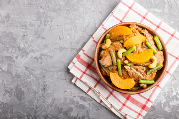 Fried pork with peaches, cashew and green beans in a wooden bowl on a gray concrete background, copy space.