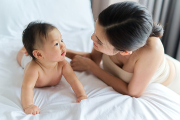 Asian cute baby crawling and playing with her beautiful mother on the bed. They are looking on each other and mother is smiling when touch the baby's back. Touch of love from mother