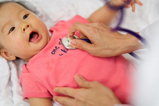 Child Doctor Is Listening Heart Pulse Rate Of Cute Newborn Baby Lying On The Bed By Using Stethoscope. Seen From Top View While The Kid Is Looking At Doctor. Baby Health Care Concept.