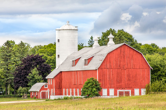 A big red wooden barn with a tall white silo stands on a farm in the rural countryside of Door County, Wisconsin.