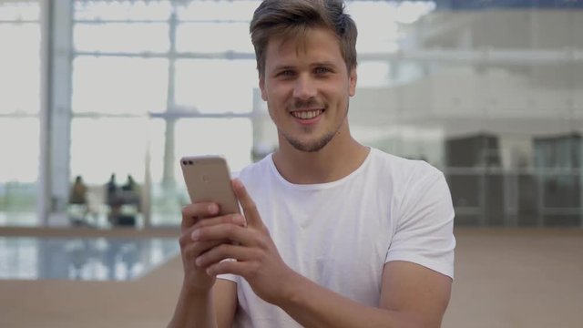 Smiling young man holding smartphone and looking at camera. Cheerful bearded guy wearing white t-shirt posing outdoor during windy day. Technology concept