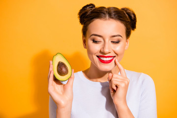 Close up photo beautiful she her lady pretty hairdo hold slice avocado look side with interest hand arm finger chin yummy tasty wear casual white pullover clothes isolated yellow background