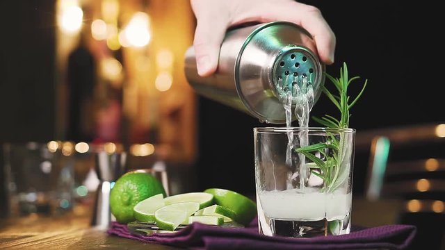 Barman's hand pouring cocktail from shaker into glass with rosemary, slow motion. Lime cocktail with rosemary on the background of the bar. Refreshing alcoholic cocktail.