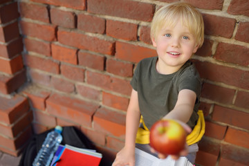 Student with big backpack and lunch bag shares his apple near the school building. School meals.