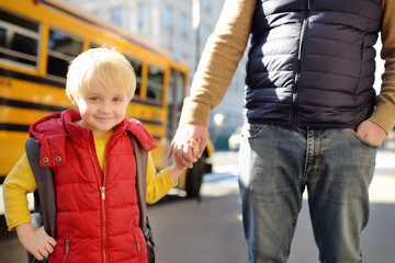 Elementary student hold hands his father near yellow school bus on background.