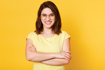 Portrait of beautiful smiling woman with folded hands isolated over yellow studio background, dressed casually, has round spectacles, has happy facial expression, being in good mood, has great news.