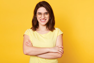 Fototapeta premium Portrait of beautiful smiling woman with folded hands isolated over yellow studio background, dressed casually, has round spectacles, has happy facial expression, being in good mood, has great news.