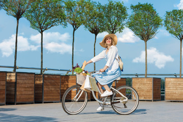 Woman is ridding around city on bicycle. Girl in blue dress and fashionable hat is going to grocery store. Vintage retro white female bicycle with basket of groceries and packages on handlebars.