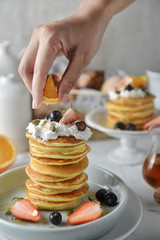 Decorating a stack of pancakes by baker hands with blueberries, strawberries, orange and whipping cream on plate.