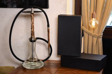 Luxury beige wooden hookah with scorched elements of lightning on stem and mouthpiece with a black hose on a brown table next to the boxes in the lounge bar on the background of a white stone wall.