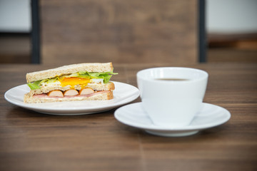 Sandwich and cup of coffee on white plate and wooden table, sandwich is made from bread, sausage, fried egg and vetgetable is American breakfast or fast food is unhealthy food or junk food