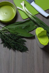 Fresh greens neatly laid out on the tabletop. Fragrant onions, dill and wild garlic. Near the container for salad, saltcellar, cutting board and kitchen knife.