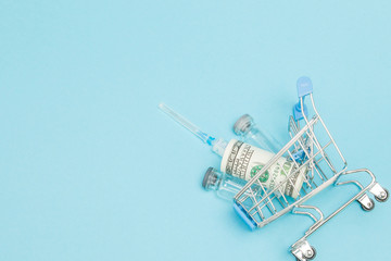 Obraz premium Medical injection and dollars in shopping trolley on blue background. Creative idea for health care cost, drugstore, health insurance and pharmaceutical company business concept. Copy space