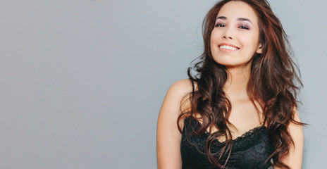 Happy sensual smiling asian young woman with dark long curly hair on grey wall background. Girl...