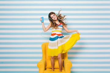 Optimistic girl wearing tn colorful dress chilling in yellow armchair and listening relaxing music and dancing with smile enjoying music. having fun on background decorated with candies.