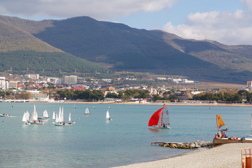 Fototapeta na wymiar The Resort Of Gelendzhik. Annual children's yacht regatta. Many yachts with white sails in Gelendzhik Bay on the background of the resort and the mountains. Markoth ridge. 
