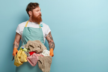 Red haired young man overstained with housekeeping, holds basin with pile of laundry, wears casual...