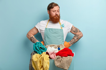 Serious thoughtful redhead man holds both hands on waist, has thick bristle, looks down, wears casual t shirt and apron, stands in front of basin with laundry and cleaning supply isolated on blue wall