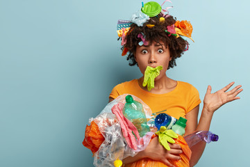 Scared frustrated Afro American woman overloaded with plastic, stuck mouth with rubber glove, has eyes popped out, worried by nature pollution, helps clean environment, free space aside for text