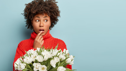 Indoor shot of surprised beautiful woman with crisp hair, covers mouth, looks aside with bated...
