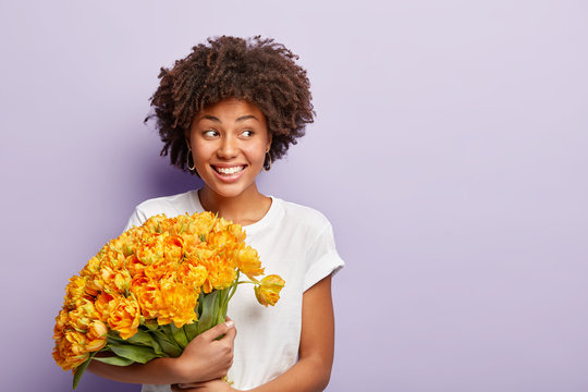Image of happy birthday woman, celebrates special day, gets big bouquet of orange flowers, wears casual t shirt, focused aside, has smile on face, looks aside, meets guests, wears casual t shirt