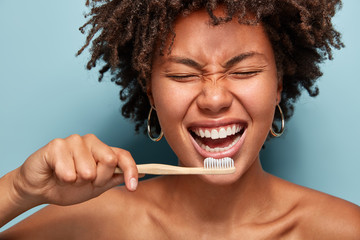 Cropped shot of cheerful glad dark skinned lady shows white teeth, has overjoyed expression, good mood in morning, prepares for dentist visit, stands with half naked body, isolated on blue wall - obrazy, fototapety, plakaty