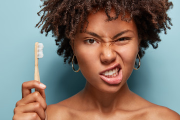 Displeased Afro woman frowns face, clenches teeth, takes care of oral hygiene, has curly hair, holds toothbrush, demonstrates bare shoulders, poses over blue background. Stomatology, whitening