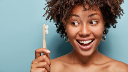 Morning rush concept. Cheerful friendly dark skinned model holds toothbrush, cares of health, wants to have healthy teeth, has brilliant smile, stands in studio over blue background with blank space