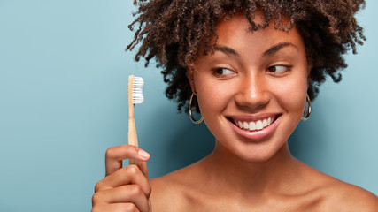 Teeth care and oral hygiene concept. Positive young Afro American woman with curly haircut, holds wooden toothbrush, cleans teeth, has dark skin, being in high spirit. Healthcare and dentistry - obrazy, fototapety, plakaty
