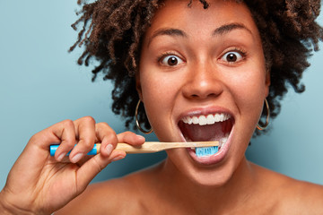 Glad mirthful woman with dark healthy skin, satisfied with new toothpaste, cleans teeth, opens mouth widely, has happy expression, curly hair, models over blue background. Close up portrait.