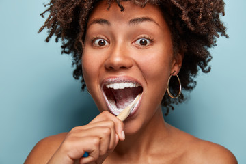 Dental care, beauty and happiness concept. Positive Afro American teen girl opens mouth widely, brushes teeth in morning with toothbrush and toothpaste, feels happy, models over blue background.