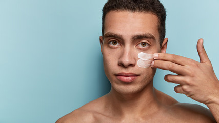 Serious male model with muscular body, applies face cream, cares of skin, stands against blue...