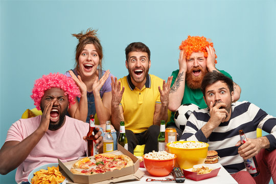 Diverse Friends Watch With Great Interest Exciting Movie, Have Surprised Expressions, Share Snack, Drink Beer, Wear Wigs, React On Interesting Scene, Isolated Over Blue Wall. People, Leisure