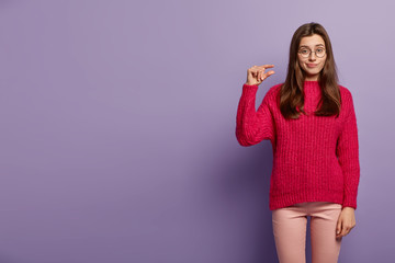 Unhappy Caucasian lady makes little small gesture, demonstrates something tiny, has unhappy expression, wears red jumper and trousers, isolated over purple background. People and size concept