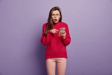 Indignant shocked young woman points at cell phone, demonstrates something amazing, surprised with received message, wears red long sleeved sweater, has astonished facial look, connected to wifi